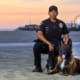 Police officer in uniform with a K9 dog on a sandy beach at sunset, pier and Ferris wheel in the background.