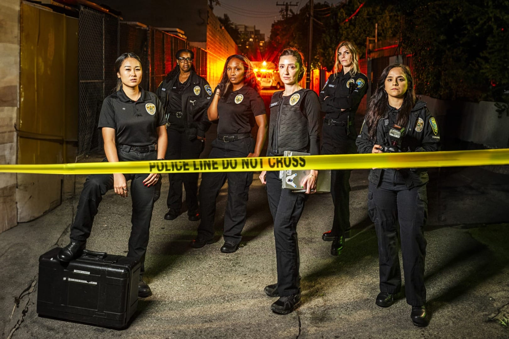Group of police officers standing behind yellow police tape in an alley, night scene with red and orange lights.