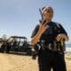 Police officer on a beach holding a radio, with a patrol vehicle and people in the background near the ocean.
