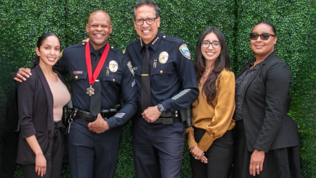 Group of five people, including two police officers, standing together and smiling in front of a green hedge background.