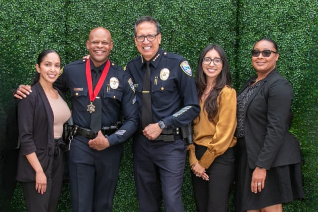 Group of five people, including two police officers, standing together and smiling in front of a green hedge background.
