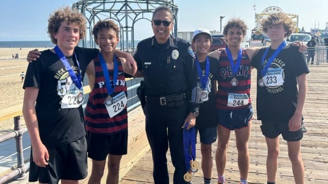 Group of five runners with medals posing with a police officer on a pier with the ocean in the background.
