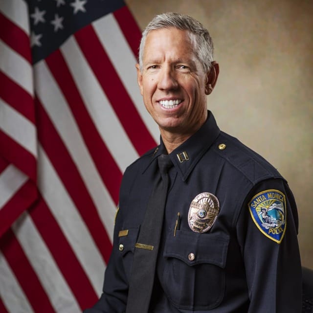 Smiling man in police uniform seated in front of American flag, wearing a badge and black tie.
