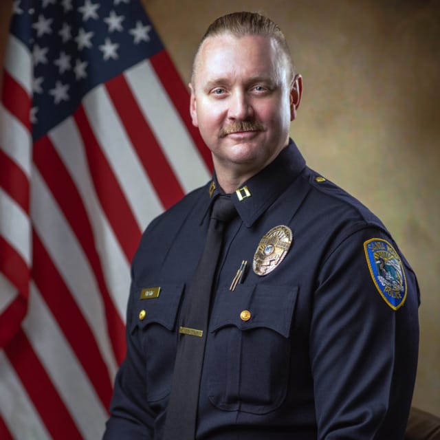 Smiling police officer in uniform seated in front of an American flag.