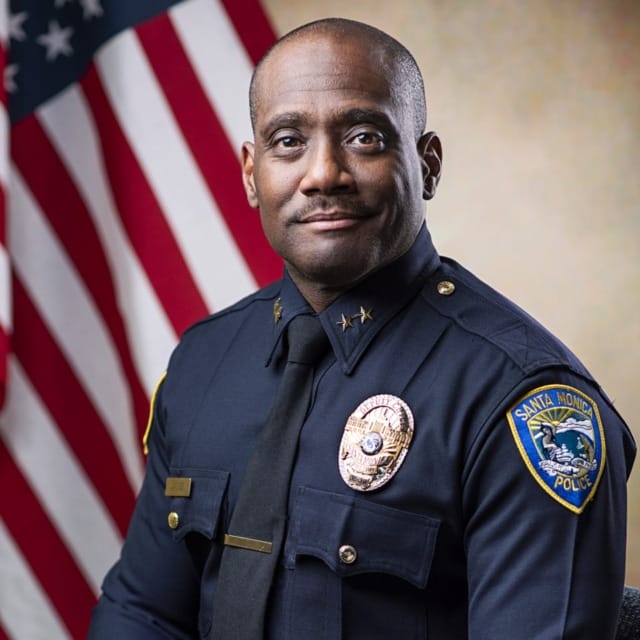 Police officer in uniform with a U.S. flag background, seated, looking forward.