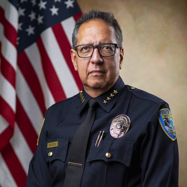 A police chief in uniform sits in front of an American flag, displaying badges and emblems.