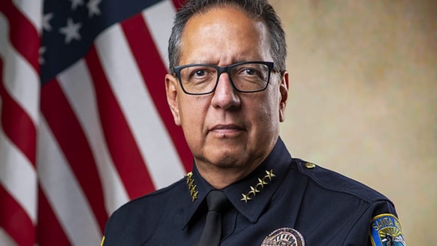 A police chief in uniform sits in front of an American flag, displaying badges and emblems.