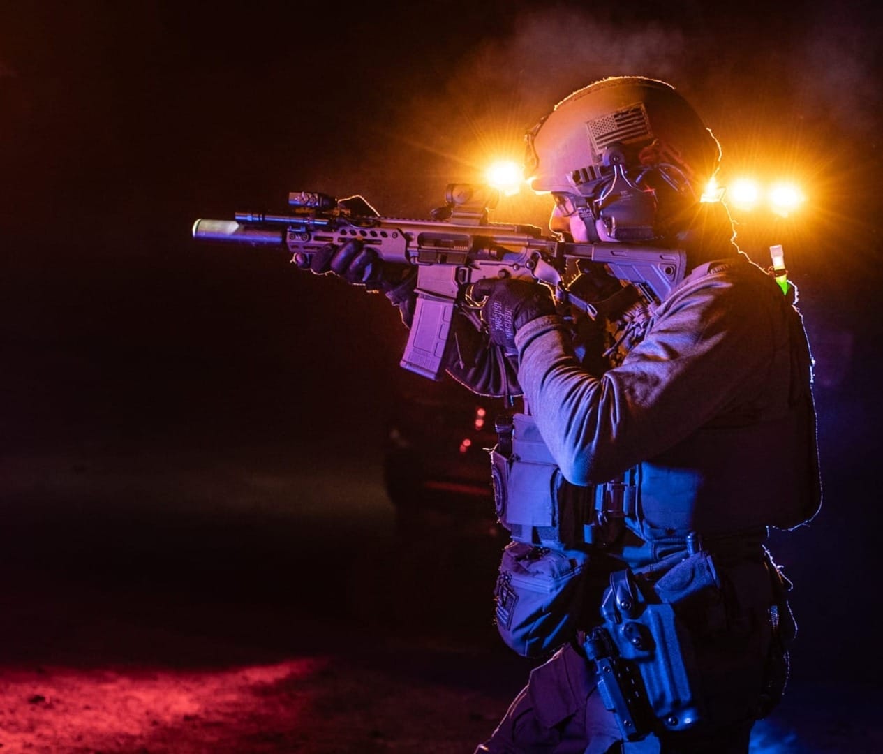 Tactical soldier aiming rifle with red and blue lights in the background at night.