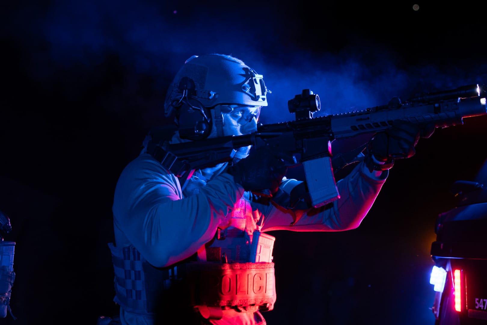 Tactical officer in gear holding a rifle, illuminated by red and blue lights, with smoke in the background at night.