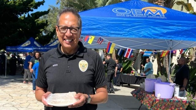 Man in uniform holds a pie at an outdoor event with tents and international flags in the background.