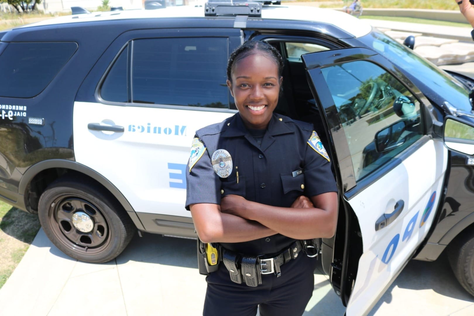Police officer standing confidently with arms crossed in front of a patrol car with an open door.