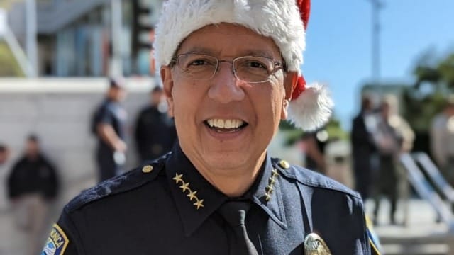 Police officer wearing Santa hat and smiling outside a building with a group in the background.