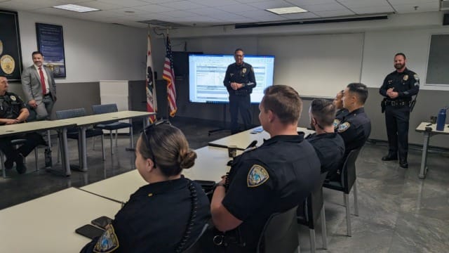 Police officers in a meeting room listening to a presentation, with a large screen displaying information in the background.