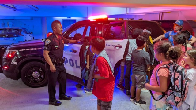 Police officer showing a police car to a group of children in a parking garage, with bright lights illuminating the scene.