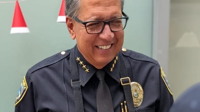 Smiling man in uniform and apron serving food, wearing a badge and glasses, with small red and white flags in the background.