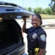 Police officer smiling and standing beside an open police vehicle trunk in an outdoor setting.