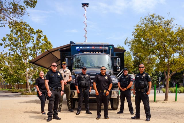 Group of police officers standing in front of a command vehicle in a park setting with trees around them.
