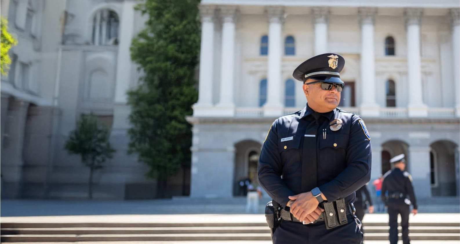 Police officer in uniform standing in front of a government building with columns, another officer in background.