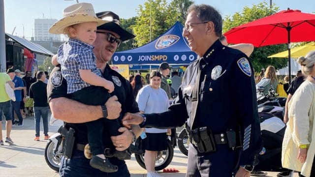 Police officers talking at an outdoor event while one holds a toddler in a cowboy hat and boots.