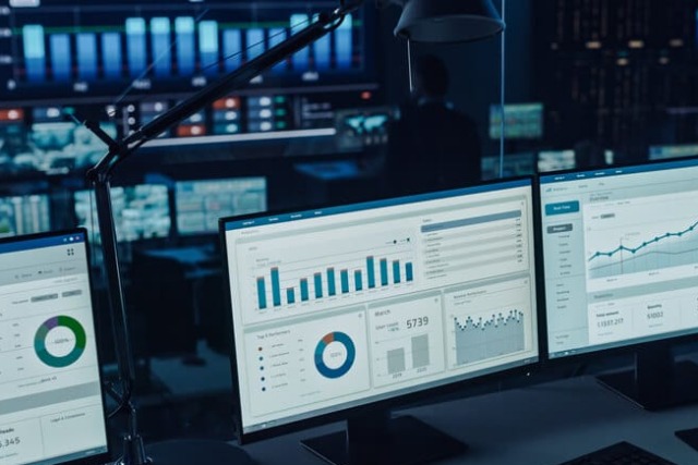 Person analyzing data on multiple computer screens in a dark office environment.