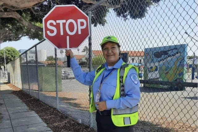 Crossing guard holding a stop sign near a fence, standing on a city sidewalk during the day.