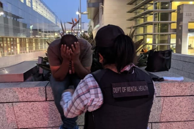 Mental health worker comforts a person in distress sitting on a wall outside a building at dusk.