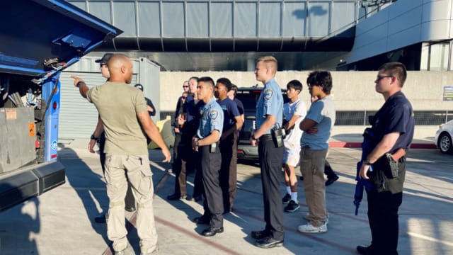 Group of people, some in uniform, standing outside listening to a man pointing toward a truck at an airport or industrial area