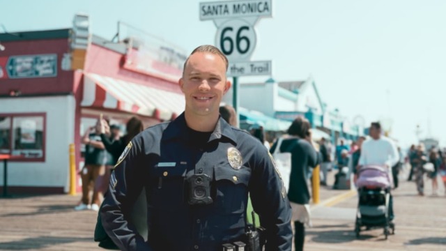 Police officer smiling at Santa Monica Pier with Route 66 sign and people walking in the background.