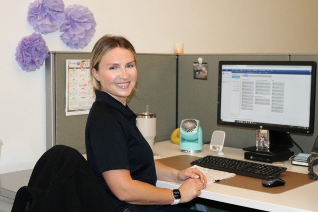 Person smiling while sitting at a desk in an office cubicle with a computer and desk fan.