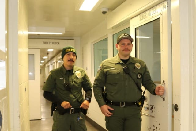 Two uniformed officers standing in a hallway, one holding a door handle labeled "Secure Holding" with a visible corridor.