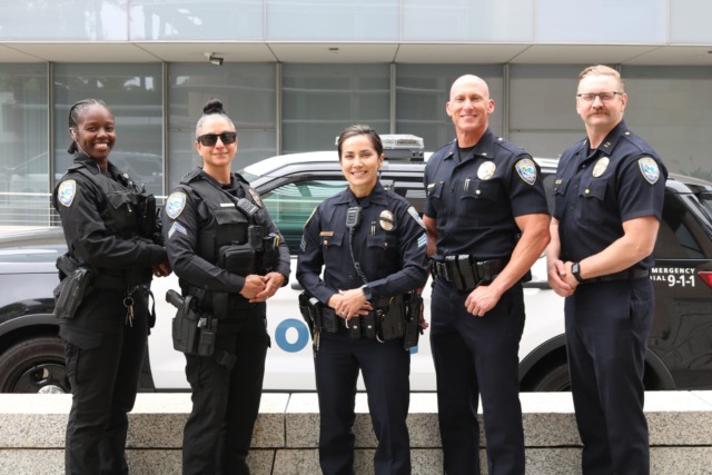 Five uniformed police officers standing in front of a patrol car, smiling at the camera.
