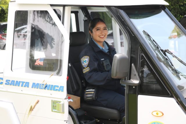 Police officer smiling inside a small vehicle, Santa Monica Police Department patch visible, American flag sticker on door.