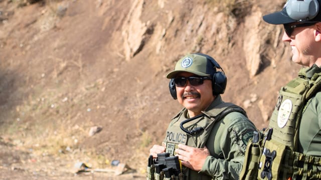 Police officers in tactical gear smiling at an outdoor shooting range with a rocky backdrop.