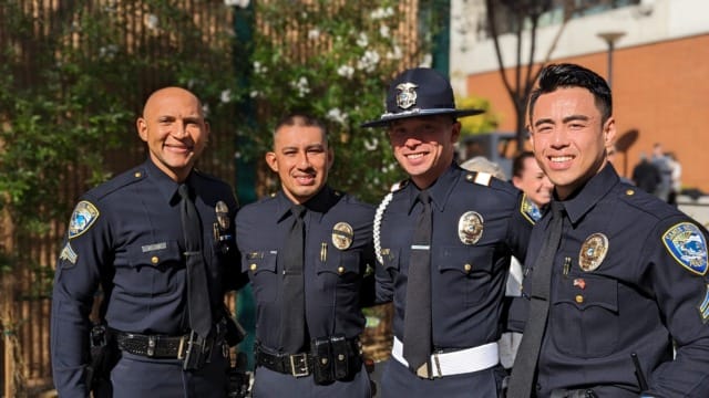 Four police officers in uniform standing together outside with trees and buildings in the background