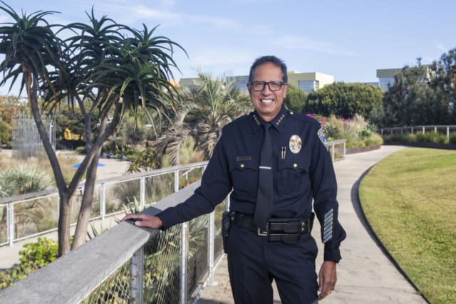 A person in a police uniform standing outdoors by a railing, with trees and buildings in the background.