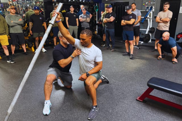 Instructor demonstrating an exercise to a group of people in a gym setting with a barbell used as a prop.