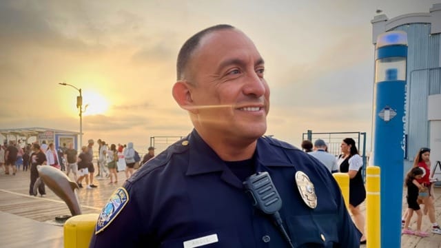 Police officer smiling on boardwalk at sunset with people in the background.