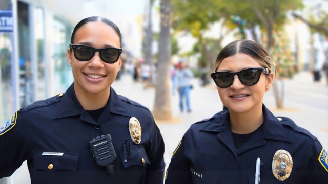 Two police officers smiling and wearing sunglasses while standing on a city street with trees and people in the background.