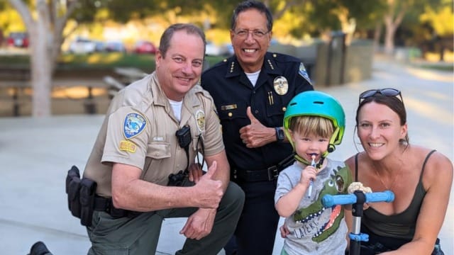 Child on scooter with helmet, posing with two officers and woman, outdoors in a park setting.