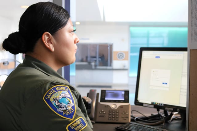 Police officer at a desk using a computer, wearing a Santa Monica police department uniform in an office setting.