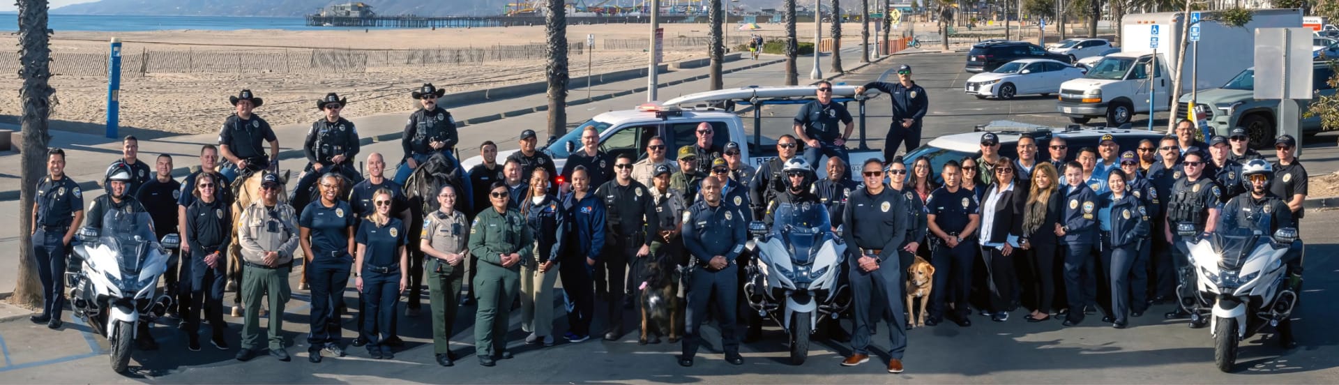 A large group of uniformed officers poses together near a beach with patrol cars and motorcycles visible in the background.