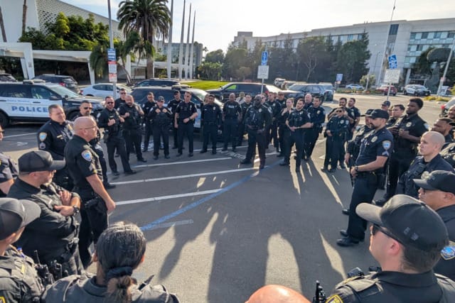 Group of police officers gathered in a parking lot having a discussion, with police vehicles parked nearby.